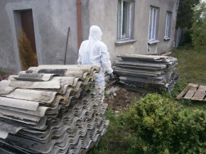 Osoba w stroju ochronnym pomiędzy dwoma pryzmami azbestu składowanymi przy budynku mieszkalnym